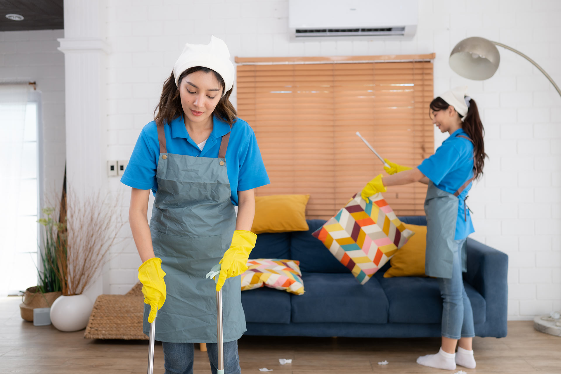 Two professional cleaners from Queen B Cleaning & Disinfecting tidy an apartment. One mops the floor while the other dusts, ensuring a fresh, clean space.