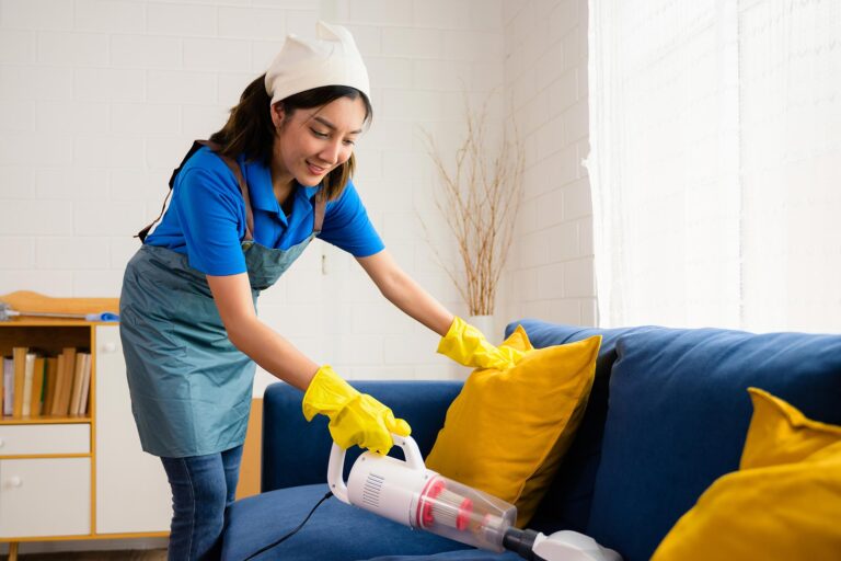 A smiling cleaner from Queen B Cleaning & Disinfecting vacuums a blue sofa with yellow cushions, providing professional apartment cleaning services.