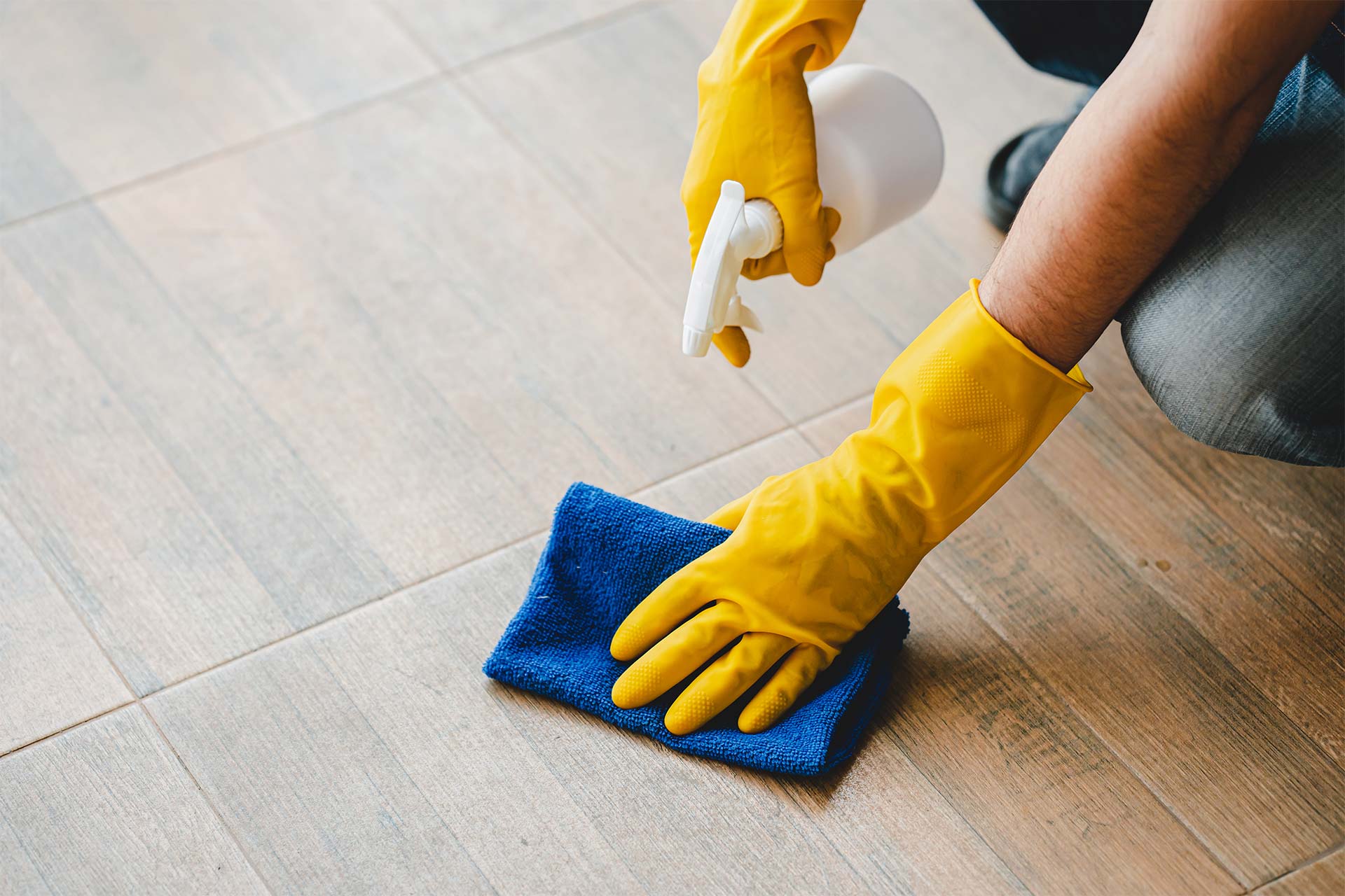 Person in yellow gloves spraying and wiping a tiled floor with a blue cloth, demonstrating professional commercial cleaning services.