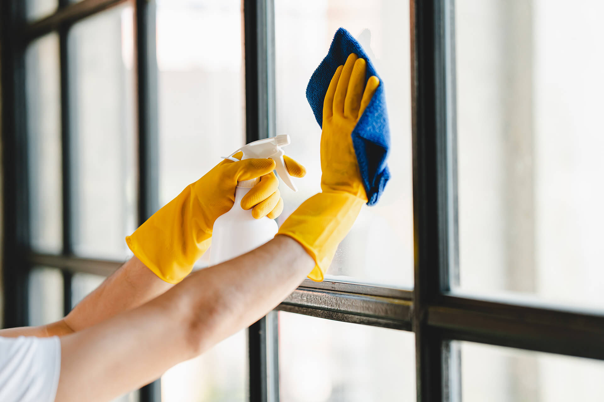 A professional cleaner from Queen B Cleaning & Disinfecting, wearing yellow gloves, meticulously cleans a commercial window with a spray and blue cloth.