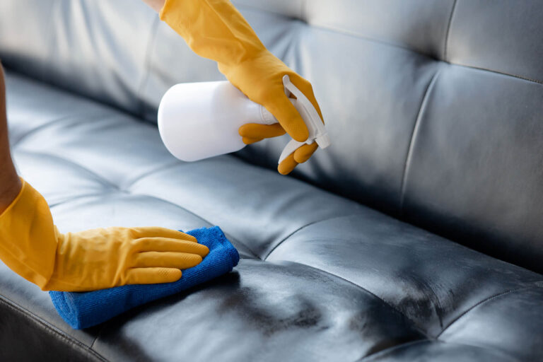 A professional cleaner in yellow gloves sprays and wipes a dark leather sofa with a blue cloth. We provide premium commercial cleaning and disinfecting services.